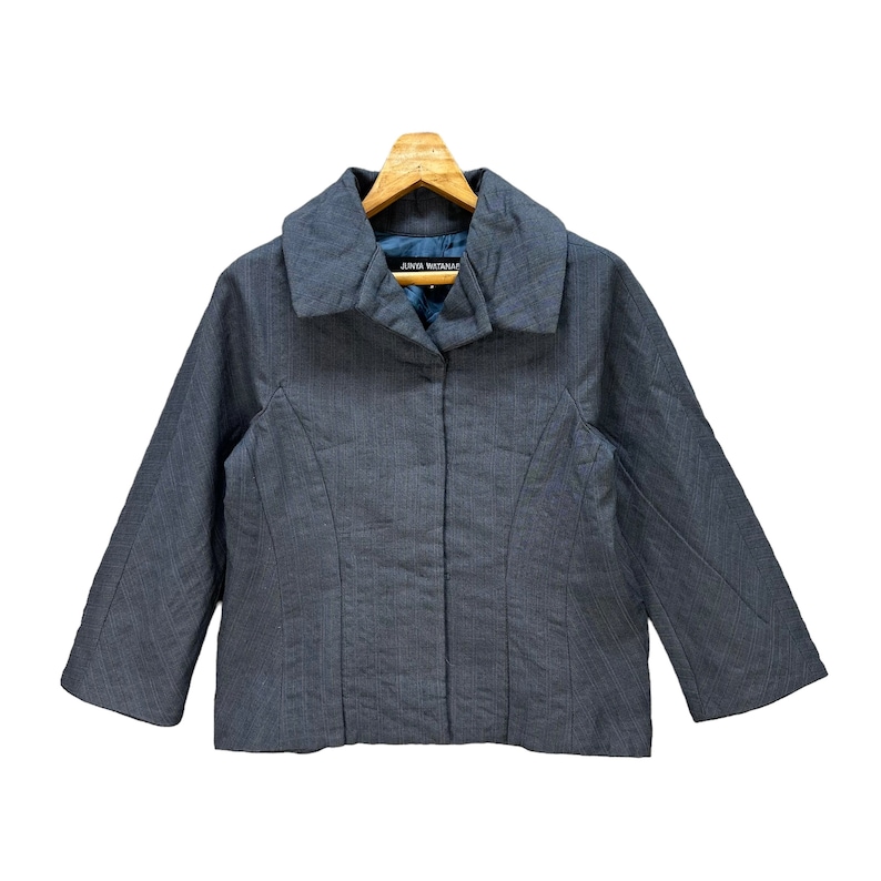 Puede incluir: Una chaqueta gris de rayas finas con cuello y cierre de botones. La chaqueta tiene mangas largas y una silueta ajustada. La etiqueta "JUNYA WATANABE" es visible en el interior del cuello.