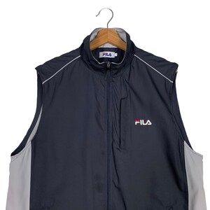 Puede incluir: Una chaqueta azul marino sin mangas con una cremallera blanca y ribetes blancos. La chaqueta tiene un panel blanco y gris en los lados y un logotipo de Fila en la parte delantera.