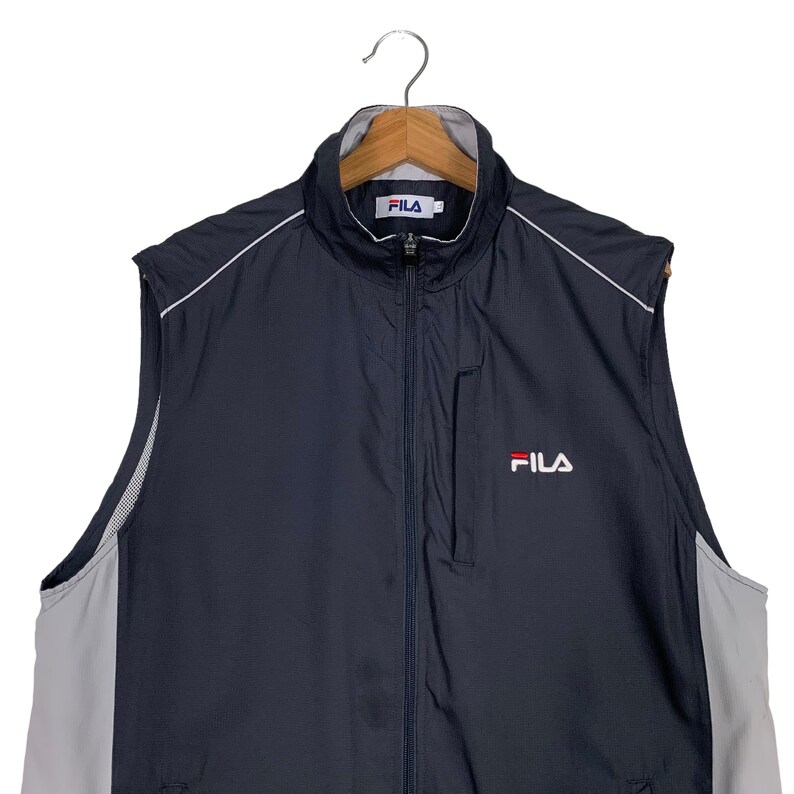 Puede incluir: Una chaqueta azul marino sin mangas con una cremallera blanca y ribetes blancos. La chaqueta tiene un panel blanco y gris en los lados y un logotipo de Fila en la parte delantera.