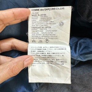 Puede incluir: Etiqueta blanca con texto negro que detalla las instrucciones de cuidado para una prenda de Comme des Gar&ccedil;ons Co., Ltd. La etiqueta dice "Made in Japan" y "Fabrique au Japon" e incluye instrucciones de lavado y secado en ingl&eacute;s y japon&eacute;s.