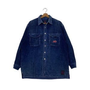 Puede incluir: Una camisa de pana azul oscuro con dos bolsillos en el pecho y una botonadura frontal. La camisa tiene cuello y mangas largas. Un pequeño logotipo rojo y blanco está bordado en el bolsillo derecho. La camisa está colgada de una percha de madera.