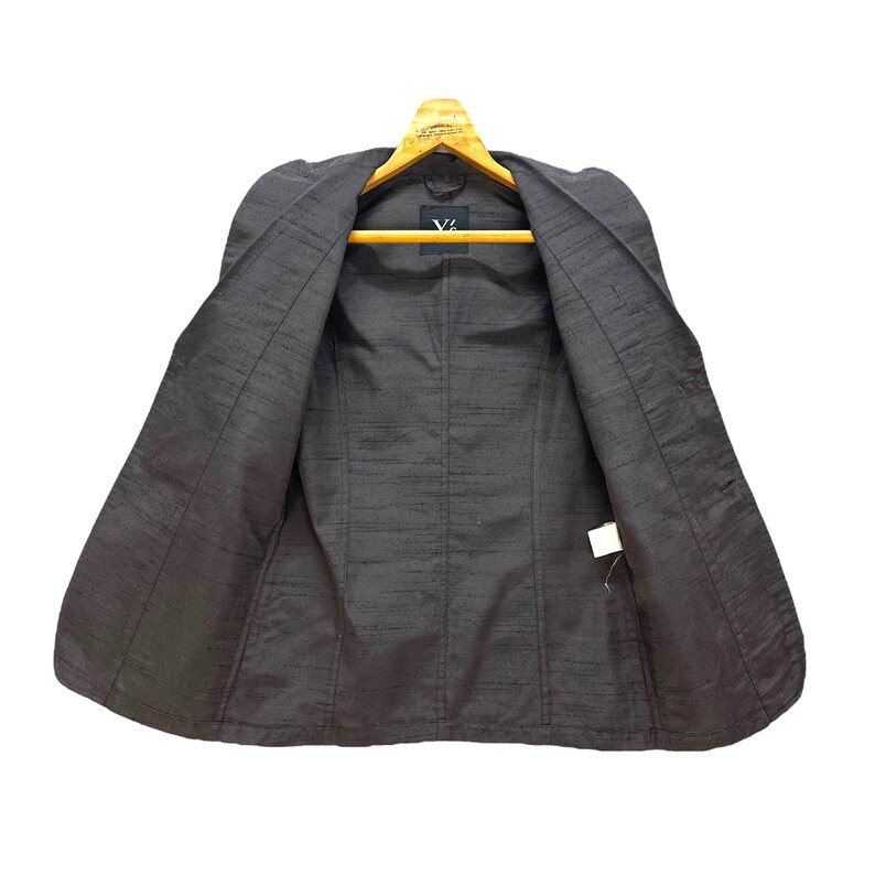 Puede incluir: Una chaqueta gris oscuro con una textura satinada. La chaqueta est&aacute; desabrochada y colocada plana sobre una superficie. El interior de la chaqueta es visible, mostrando un forro gris oscuro.