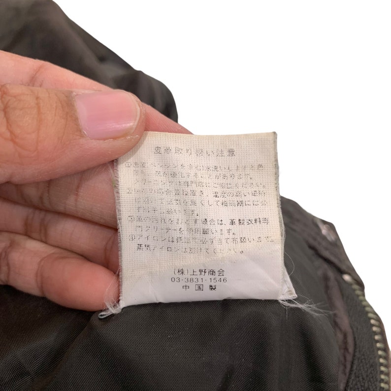 May include: A white tag with black text detailing care instructions for leather goods. The tag reads "Leather Care Instructions" and includes information about washing, drying, and ironing. The tag also includes the company name "(株)上野商会" and the phone number "03-3831-1546". The tag is made in China.
