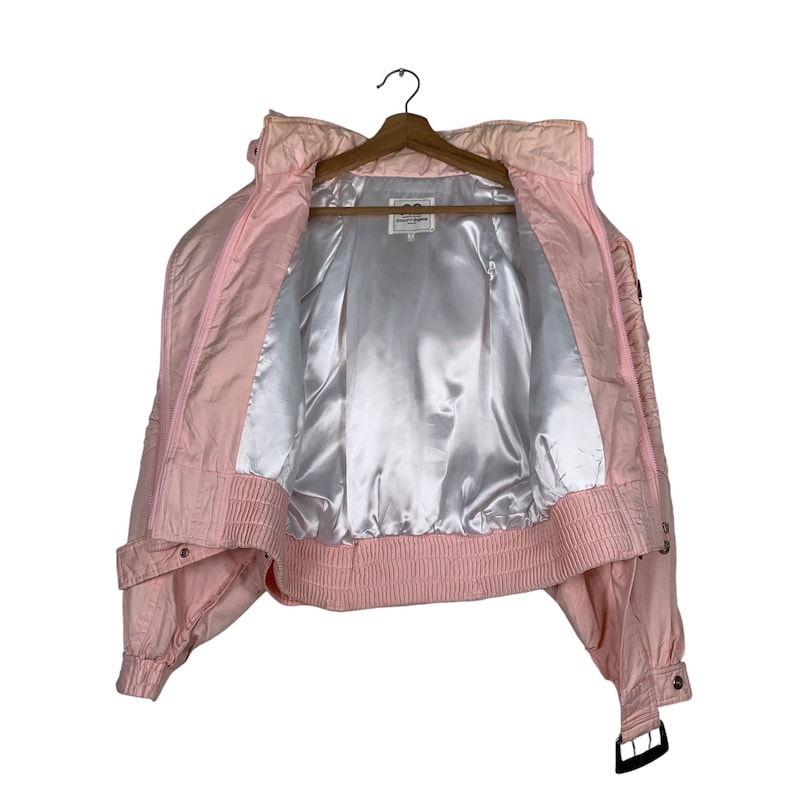 May include: A pink puffer jacket with a satin lining and a zip closure. The jacket has a cropped fit and a belt at the waist.