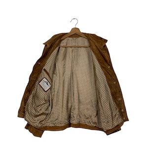 May include: A brown suede jacket with a collared neckline and button closures. The jacket is open, revealing a patterned lining and a small label with a logo. The jacket is hanging on a wooden hanger.