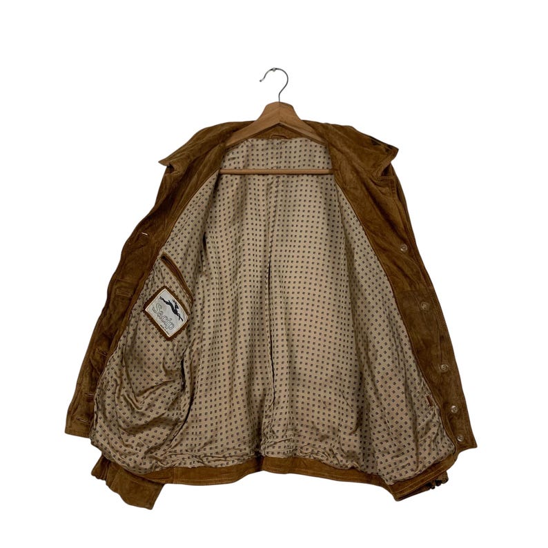 May include: A brown suede jacket with a collared neckline and button closures. The jacket is open, revealing a patterned lining and a small label with a logo. The jacket is hanging on a wooden hanger.