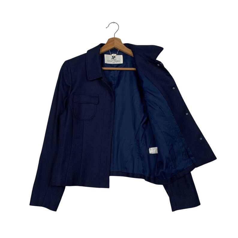 May include: A navy blue jacket with a collared neckline and a single pocket on the left side. The jacket is open, revealing a dark blue interior lining. The jacket is hanging on a wooden hanger.