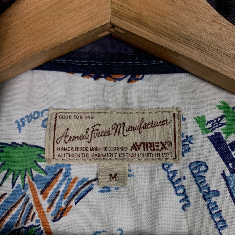 May include: A white shirt with a blue and green palm tree print. The shirt has a brown label that reads "Good for One Armed Forces Manufacturer AVIREX&reg; Name & Trade Mark Registered Authentic Garment Established in 1975". The label also has a size "M" tag.