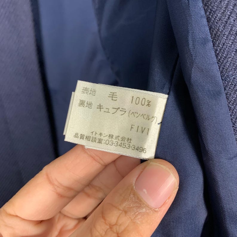 May include: Close-up of a tag with Japanese text and English text. The tag is white with black text and is held against a blue fabric. The text includes "表地 毛 100%" and "FIVI".