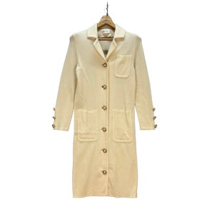 May include: A long, cream-colored knitted cardigan with a button-up front and two large pockets. The cardigan has gold-toned buttons and decorative accents on the cuffs.
