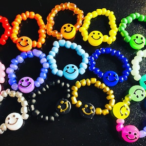 Pode incluir: Uma coleção de anéis de contas coloridas com pingentes de carinhas sorridentes. Os anéis são feitos com contas de diferentes cores e apresentam uma variedade de pingentes de carinhas sorridentes de diferentes cores.