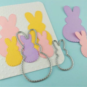 May include: A set of five die-cut Easter bunny shapes in pink, yellow, and purple. The bunnies are layered on top of each other, with the largest bunny on the bottom and the smallest bunny on top. The bunnies are all cut out of cardstock and have a stitched edge.