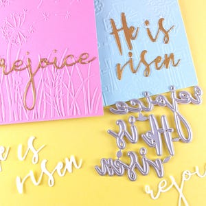 May include: Pink and blue Easter-themed greeting cards with embossed floral designs and the phrases "rejoice" and "He is risen" in gold and white script. Die-cut metal words are also present.