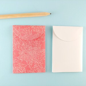 May include: A coral card with a white floral pattern and a white envelope are placed on a light blue surface. A wooden pencil is in the top left corner. The scene is simple and elegant.