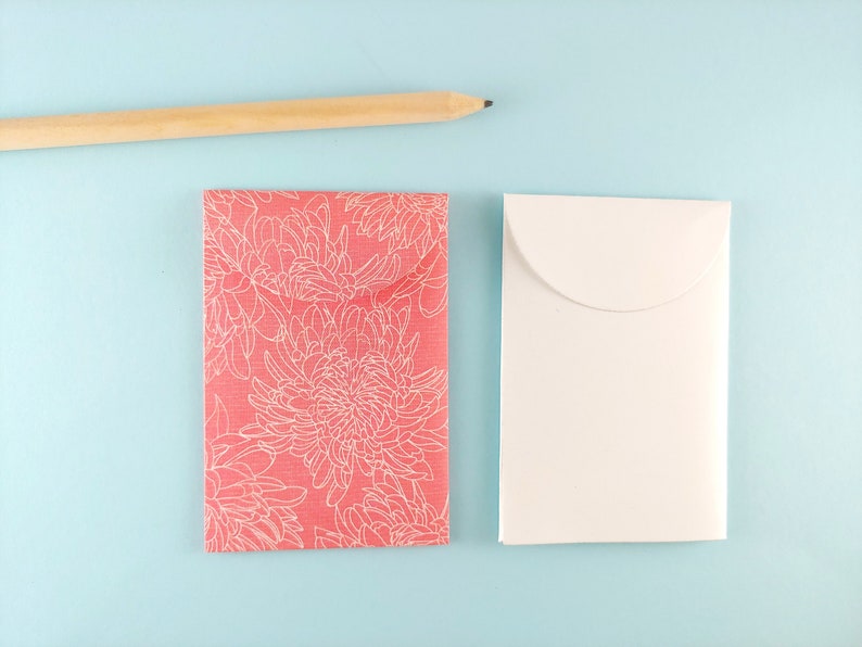 May include: A coral card with a white floral pattern and a white envelope are placed on a light blue surface. A wooden pencil is in the top left corner. The scene is simple and elegant.