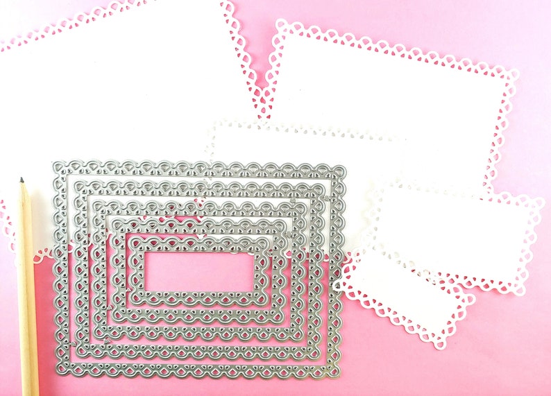 May include: A set of six silver metal die-cut frames with intricate scalloped edges. The frames are arranged in a nested pattern, with the largest frame at the bottom and the smallest frame at the top. The frames are on a pink background.
