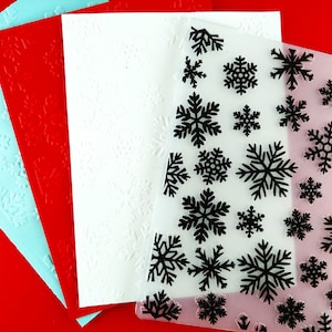 Puede incluir: Cuatro hojas de papel con diseños de copos de nieve. La hoja superior es azul claro, la segunda hoja es roja, la tercera hoja es blanca y la hoja inferior es transparente con diseños de copos de nieve negros.