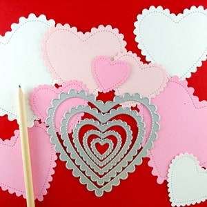 May include: A collection of pink and white heart-shaped paper cutouts with scalloped edges and a pencil on a red background. Several nested heart-shaped metal dies are also present, suggesting a crafting theme. The hearts vary in size.