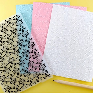 May include: Three sheets of clear plastic with an embossed floral pattern. The sheets are in different colors: light blue, pink, and white. A pencil is lying on the table next to the sheets.