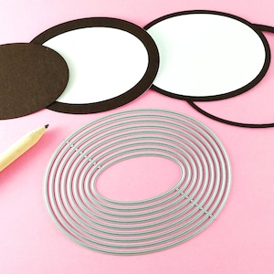 May include: A set of oval-shaped metal craft dies with multiple layers, arranged in a concentric pattern on a pink surface. A brown paper circle, a white paper circle, and a brown paper ring are also visible.