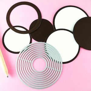 May include: A set of craft supplies for creating layered circles. The set includes a silver metal die with multiple concentric circles, brown cardstock circles, and white cardstock circles.