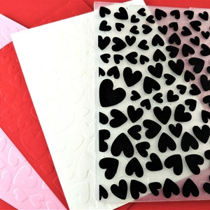 May include: Four sheets of paper with embossed heart patterns. The sheets are pink, red, white, and clear with black hearts. The clear sheet is a stencil for crafting.