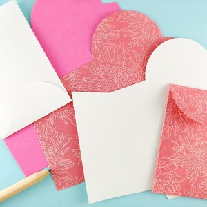 May include: A collection of stationery items, including white and pink envelopes, and cards. Some envelopes are solid pink, while others feature a coral floral pattern. A wooden pencil rests on the light blue surface, near the stationery.