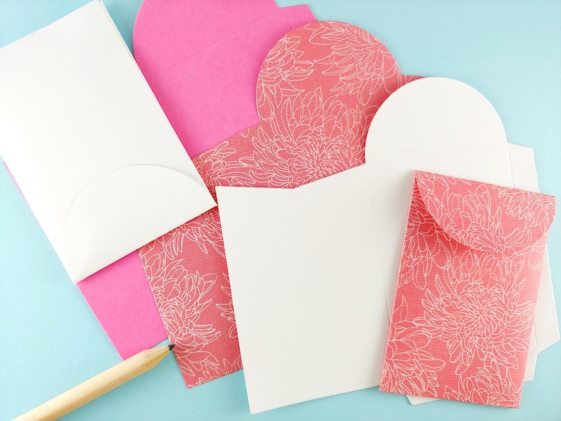 May include: A collection of stationery items, including white and pink envelopes, and cards. Some envelopes are solid pink, while others feature a coral floral pattern. A wooden pencil rests on the light blue surface, near the stationery.