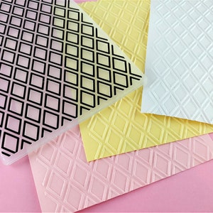 May include: A clear plastic embossing plate with a black diamond pattern. Three sheets of paper are embossed with the same pattern in pink, yellow, and white.