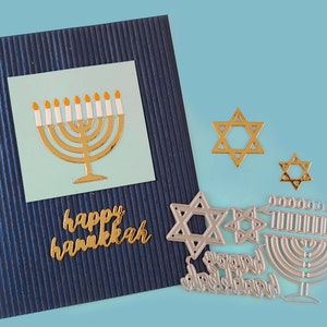 May include: A blue card with gold lettering that says "happy hanukkah" and a gold menorah with white candles on a light blue background. There are also several silver metal die cuts of menorahs and Stars of David.