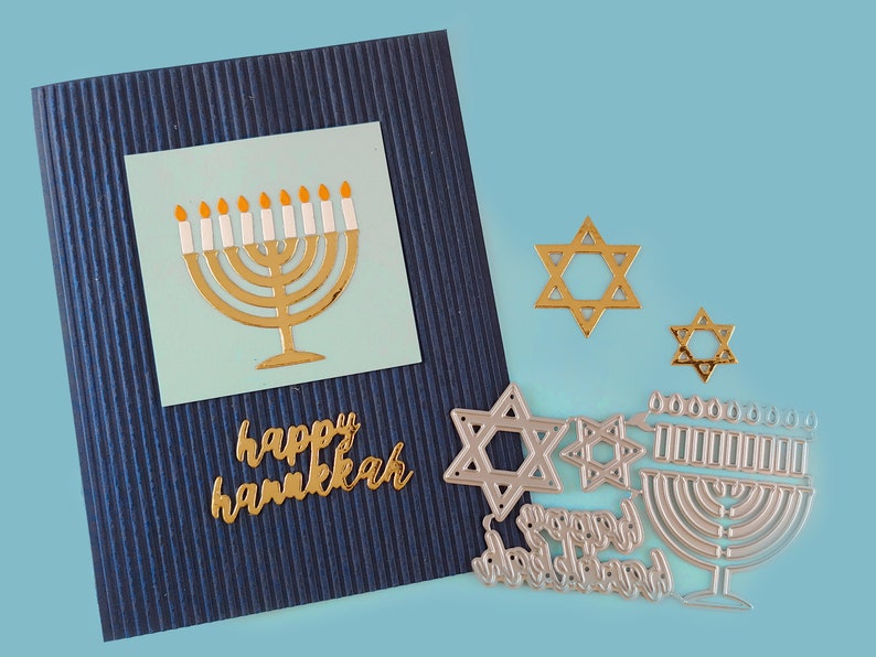 May include: A blue card with gold lettering that says "happy hanukkah" and a gold menorah with white candles on a light blue background. There are also several silver metal die cuts of menorahs and Stars of David.