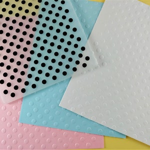 May include: Four sheets of paper with a polka dot pattern. The top sheet is clear with black polka dots. The next sheet is light blue with white raised polka dots. The third sheet is white with white raised polka dots. The bottom sheet is pink with white raised polka dots.