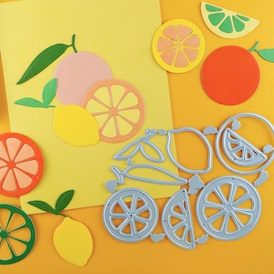 May include: A set of metal craft dies for cutting out citrus fruit shapes, including lemons, limes, and oranges. The dies are arranged on a yellow surface with paper cutouts of citrus fruits.