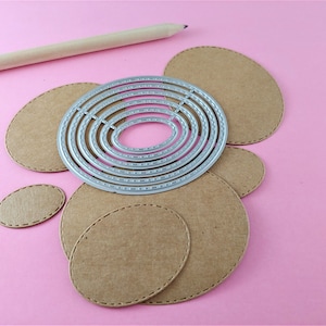 May include: A set of six metal oval-shaped die-cut stencils with a decorative stitched edge. The stencils are arranged in a circle on a pink surface with brown paper circles cut with the stencils.