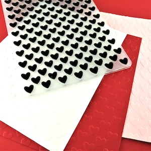 May include: Three sheets of paper with a heart pattern embossed on them. The top sheet is clear plastic with black hearts, the middle sheet is white with white hearts, and the bottom sheet is red with red hearts.
