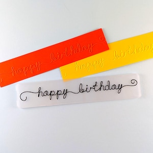 May include: Three rectangular, colorful birthday embellishments. One is orange, another is yellow, and the third is white. Each has the words "happy birthday" in a cursive font. The orange and yellow rectangles have the words embossed, while the white one has the words in black.