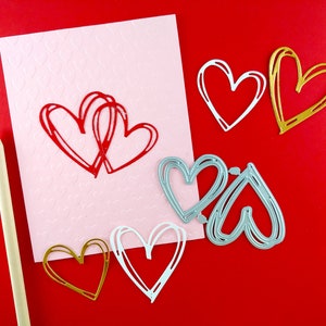 May include: A selection of heart-shaped craft materials against a red backdrop. Features a pink card with embossed hearts and red drawn hearts, alongside gold, white, and silver heart-shaped cutouts. A pencil is also included.