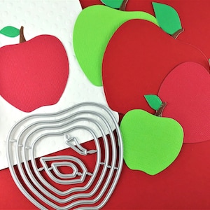 May include: A set of five apple-shaped die-cut craft supplies. The set includes two red apples, two green apples, and a silver metal die-cut apple shape. The apples are on a red background.