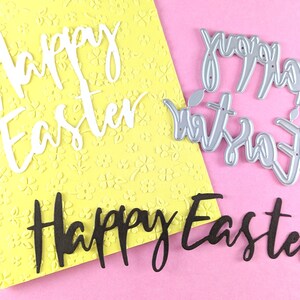 May include: A close-up of Easter-themed craft supplies. The image features the words "Happy Easter" in white, black, and silver, with a yellow floral background and a pink surface. The craft supplies include die cuts and a card.