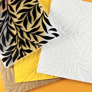 May include: A set of three leaf patterned embossing plates. The top plate is clear with black leaves, the middle plate is yellow with white leaves, and the bottom plate is white with raised white leaves.