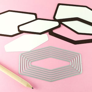 May include: A set of six die-cut hexagon shapes in brown and white, and a set of seven silver metal hexagon dies. The dies are arranged on a pink surface.