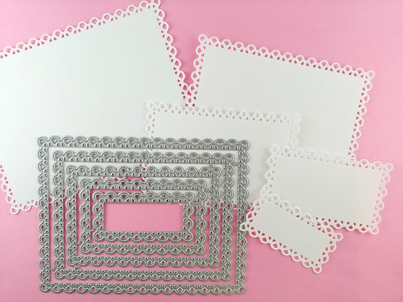 May include: A set of six silver metal die-cut frames with scalloped edges. The frames are arranged in decreasing size, with the largest frame at the top and the smallest frame at the bottom. The frames are on a pink background.