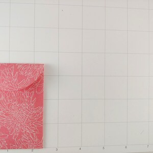 May include: A small, rectangular, pink envelope with a white floral pattern. The envelope is folded and has a flap. The background is a white grid.