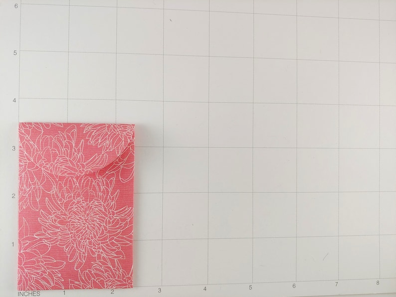 May include: A small, rectangular, pink envelope with a white floral pattern. The envelope is folded and has a flap. The background is a white grid.