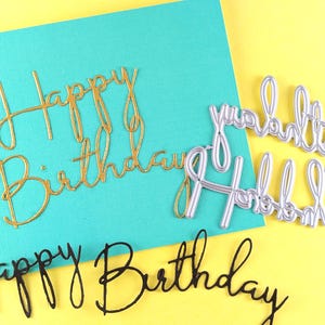 May include: A collection of die-cut metal words spelling "Happy Birthday". The words are in gold, silver, and black, with a teal card and yellow background. The script font is elegant and suitable for crafting.