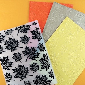 May include: Four sheets of paper with an embossed maple leaf pattern. The sheets are in orange, grey, yellow, and clear with black maple leaves.
