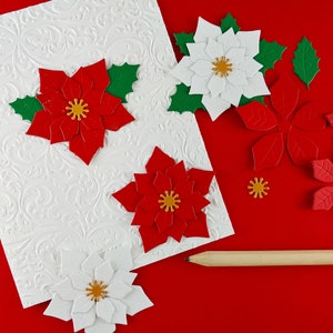 May include: A white card with a textured pattern is decorated with red and white paper poinsettia flowers. Green paper leaves and a wooden pencil are also included.