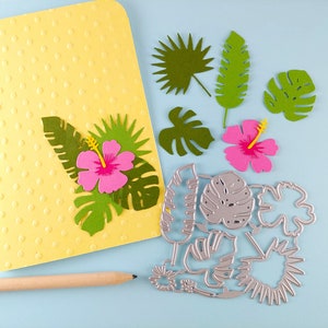 May include: A set of tropical leaf and flower die cuts in green, pink, and yellow. The die cuts are arranged on a light blue surface with a yellow card and a wooden pencil.