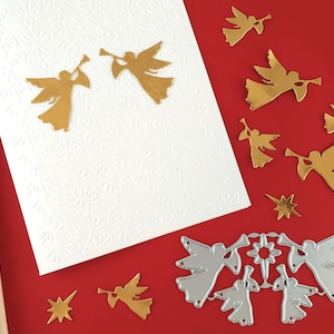 Peut inclure: Une carte blanche avec un motif gaufré doré et deux découpes d'anges dorées. La carte est sur une surface rouge avec plusieurs découpes d'anges dorées et une découpe d'étoile argentée. Il y a aussi une découpe d'ange en argent avec une étoile au centre.