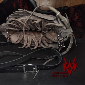 May include: A distinctive grey leather handbag with a unique design. It features layered, textured elements and a long, adjustable black strap. The bag has a silver-tone buckle and hardware, and a top handle.